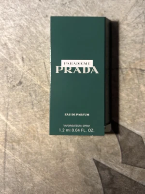 Prada Paradigme Eau de Parfum 1,2 ml - Prada Paradigme Eau de Parfum i en elegant mörkgrön kartong med vit text. Flaskan är liten och transparent med sprayfunktion, perfekt för att testa doften eller ha med sig i väskan. Volymen är 1,2 ml.