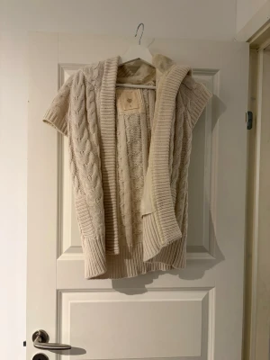 Beige stickad väst med luva - Säljer en beige stickad väst med luva och korta ärmar. Västen har ett klassiskt flätmönster och breda ribbade muddar. Perfekt att slänga över en långärmad tröja för en chill och snygg look. Priset är inte hugget i sten vid snabb affär!