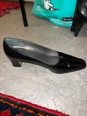 Svarta pumps med låg klack - Stilrena svarta pumps från Schell i blankt skinnmaterial. Skorna har låg, bred klack och en fyrkantig tå för en klassisk look. Insidan är grå med diskreta prickdetaljer på innersulan. Perfekta för dig som gillar tidlös och enkel stil.