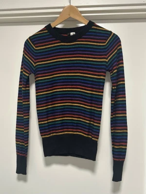 Svart stickad H&M tröja med regnbågsränder - Finstickad tröja från H&M Divided i storlek S / 34 med långa ärmar och rund halsringning. Tröjan har ett randigt mönster i svart, röd, orange, gul, grön, blå och lila. Mycket gott skick, helt noppfri.   Skickas mot porto, kan även hämtas i Stockholm.