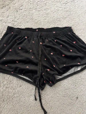 Svarta pyjamasshorts med hjärtan - Mysiga svarta pyjamasshorts i mjukt velourmaterial med små rosa hjärtan över hela. Shortsen har resår i midjan och dragsko för extra komfort. Perfekta för att chilla hemma eller sova i.