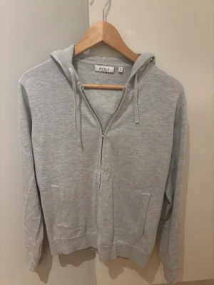 Ljusgrå zip hoodie från RYVLS - Ljusgrå hoodie med dragkedja från RYVLS. Klassisk modell med huva och dragsnören. Tillverkad i mjukt sweatshirtmaterial, perfekt för en avslappnad stil. Storleken är 158-164 och modellen är 175 cm lång.