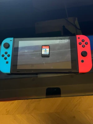 Nintendo Switch med blå och röd Joy-Con - Nintendo Switch spelkonsol med en blå Joy-Con till vänster och en röd Joy-Con till höger. Konsolen har en stor skärm i mitten och är tillverkad i plast. Perfekt för gaming både hemma och på språng. Ett spelkort syns på skärmen.