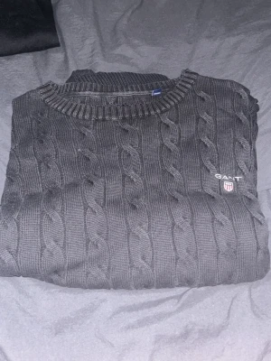 Svart kabelstickad tröja från GANT - Kabelstickad svart tröja från GANT i premium bomull. Klassisk rund halsringning och diskret GANT-logga broderad på bröstet. Snygg ribbad kant vid halsen och ett stilrent mönster som ger en tidlös look.