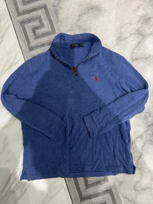 Blå halvzip tröja från Polo Ralph Lauren - Snygg blå långärmad tröja från Polo Ralph Lauren med halvzip och klassisk röd logga broderad på bröstet. Tröjan har hög krage och är tillverkad i mjukt material, perfekt för lager-på-lager. Passar dig som gillar stilren design.