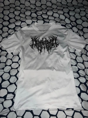 Vit t-shirt med svart tryck - Säljer en vit t-shirt med ett stort svart grafiskt tryck på bröstet. T-shirten har rund halsringning och korta ärmar. Materialet känns mjukt och skönt, troligtvis bomull. Perfekt för dig som gillar streetwear och vill sticka ut.