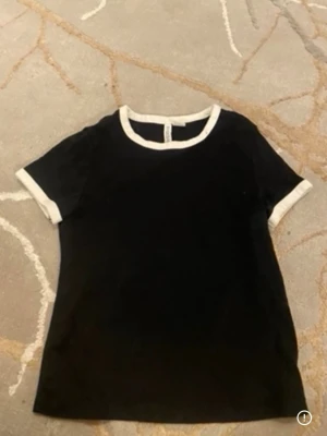 Svart t-shirt med vita detaljer - En stilren svart t-shirt med vita kanter vid halsringning och ärmslut. Klassisk passform och rund hals. Perfekt till jeans eller kjol för en enkel och snygg look.