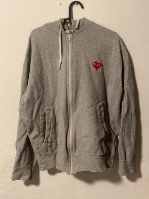 Grå hoodie från Comme des Garçons Play - Grå zip-hoodie från Comme des Garçons Play med huva och dragsko. Fram finns ett rött hjärta med ögon och på ryggen flera röda hjärtan samt PLAY-tryck. Tillverkad i mjuk bomullsblandning och har två fickor framtill. Perfekt för en chill och trendig look.
