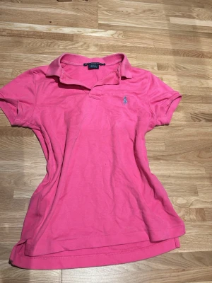 Rosa pikétröja från Ralph Lauren - Säljer en klassisk rosa pikétröja från Ralph Lauren Sport i slim fit-modell. Tröjan har korta ärmar, krage och knappar vid halsen samt den ikoniska broderade loggan på bröstet. Tillverkad i mjuk bomull som är skön att bära.
