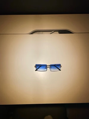 Blå rektangulära solglasögon - Snygga solglasögon med rektangulära, blåtonade glas och tunn metallbåge i silver. Glasen är utan ram runt och har en modern, minimalistisk look. Perfekta för dig som vill sticka ut med en trendig accessoar.