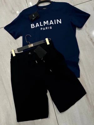 Balmain Paris set i blått och svart - Snyggt set från Balmain Paris med en mörkblå t-shirt i bomull och vit logga framtill samt matchande svarta shorts med dragsko och liten logga. Perfekt för en trendig och avslappnad look. Materialet känns mjukt och skönt mot huden.