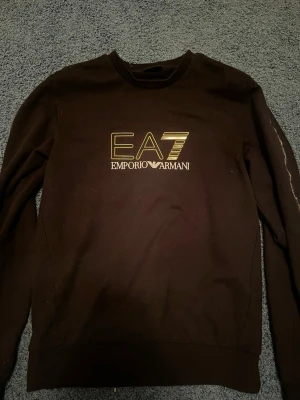 Svart EA7 Emporio Armani sweatshirt - Snygg svart sweatshirt från EA7 Emporio Armani med guldig logga framtill. Tröjan har rund halsringning och långa ärmar. Perfekt för dig som gillar stilrena och exklusiva plagg. Materialet känns mjukt och bekvämt mot huden.