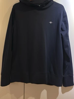 Svart hoodie från GANT med logga - Snygg svart hoodie från GANT med liten broderad logga på bröstet. Klassisk modell med huva och ribbade muddar. Tillverkad i mjukt och bekvämt material som passar perfekt till vardagslooken. Enkel och stilren design som funkar till det mesta.