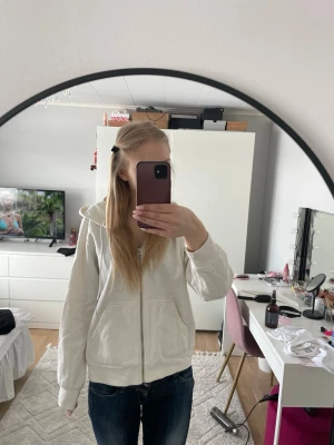 Vit hoodie med dragkedja - Säljer en vit hoodie med dragkedja och huva. Modellen har ribbade muddar vid ärmslut och nederkant samt två fickor fram. Perfekt för en avslappnad och chill look. Materialet ser ut att vara mjukt och bekvämt, troligen bomullsblandning.