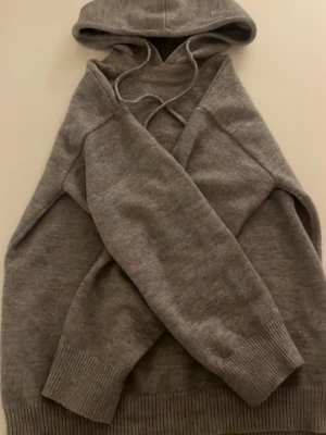 Grå hoodie i kashmire - En stilren grå hoodie med huva och dragsnören. Tillverkad i mjuk stickad kvalitet med breda ribbade muddar vid ärmslut och nederkant. Perfekt för en avslappnad look och enkel att matcha med jeans eller mjukisbyxor.