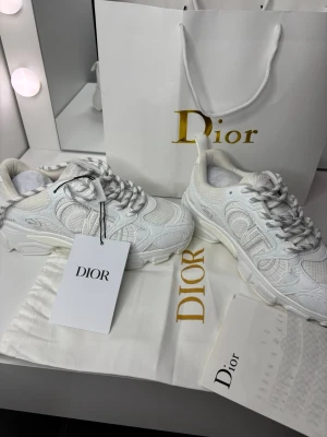 Vita  sneakers med mönster - Säljer ett par vita Dior sneakers med snygga detaljer och mönster i ton-i-ton. Skorna har tjock sula, broderade partier och klassiska snörningar. Märkeslogga syns på sidan och tungan. Perfekta för dig som vill sticka ut med exklusiva sneakers.