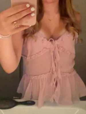 Ljusrosa volang topp - Supersöt ljusrosa topp med volangdetaljer och snörning framtill. Toppen är ärmlös och har en feminin, romantisk look med v-ringning och lätt transparent material. Perfekt att styla med jeans eller kjol för en trendig outfit.