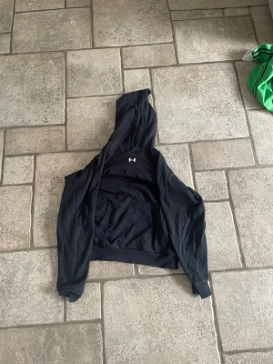 Svart hoodie från Under Armour - Säljer en svart hoodie från Under Armour med klassisk logga på bröstet. Hoodien har huva med dragsko och en stor magficka. Tillverkad i mjukt material som känns skönt mot huden. Perfekt för chill dagar eller träning.