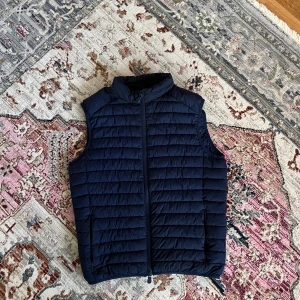 Mörkblå dunväst från Save The Duck - Snygg mörkblå dunväst från Save The Duck i ultralätt syntetmaterial. Västen har horisontella quiltade sömmar, hög krage och dragkedja framtill. Perfekt för lager-på-lager och enkel att matcha med olika outfits.