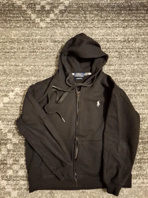 Svart ziphoodie från Polo Ralph Lauren - Klassisk svart hoodie från Polo Ralph Lauren med dragkedja framtill och vit broderad logga på bröstet. Modellen är custom fit och har justerbar huva med snören samt ribbade muddar vid ärmslut och nederkant. Perfekt för en avslappnad och stilren look.