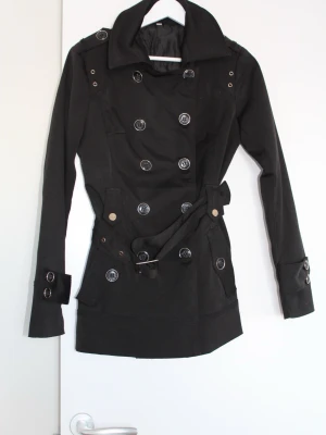 Svart trenchcoat med bälte och knappar - Stilren svart trenchcoat med dubbelknäppning, stora silvriga knappar och bälte i midjan. Jackan har bred krage, dekorativa spännen vid ärmslut och fickor med knappdetaljer. Fodrad insida för extra komfort. Perfekt för en trendig look.