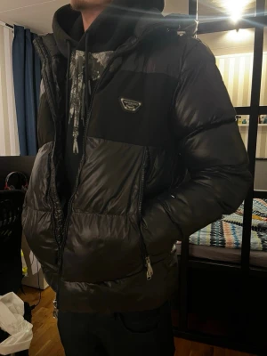 Alessandro Zavetti Jacka - Snygg svart pufferjacka från Alessandro med huva och dragkedja. Jackan har en logotyp på bröstet och stor broderad text på ryggen. Fylld för extra värme och har fickor med dragkedja. Perfekt för kalla dagar och riktigt streetig look.