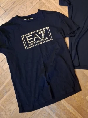 Svart EA7 Emporio Armani t-shirt - Snygg svart t-shirt från EA7 Emporio Armani med guldig logga framtill. Klassisk rund halsringning och korta ärmar. Perfekt för dig som gillar stilrena märkesplagg.