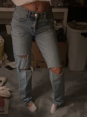 Ljusblå medelhöga jeans - Säljer ett par ljusblå jeans med raka ben och slitna detaljer vid knäna. Jeansen har klassisk femficksdesign och hög midja. Perfekta för en avslappnad och trendig look.