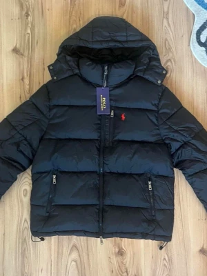 Svart pufferjacka från Polo Ralph Lauren - Svart pufferjacka från Polo Ralph Lauren med röd logga på bröstet. Jackan har huva, dragkedja framtill och två fickor med dragkedja. Klassisk quiltad design och varm vaddering, perfekt för kalla dagar.