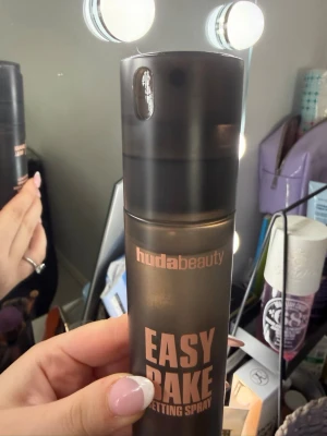 Huda Beauty Easy Bake Setting Spray - Face mist från Huda Beauty i svart, matt plastflaska med roséguldfärgad text. Easy Bake Setting Spray ger en fräsch finish och håller sminket på plats hela dagen. Praktisk sprayfunktion och stilren design som passar perfekt på sminkbordet.