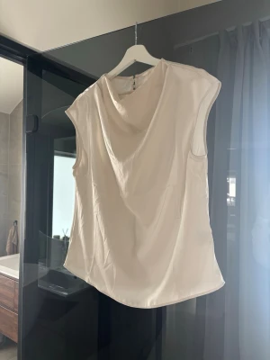 Vit ärmlös blus med draperad hals - Stilren vit blus med draperad halsringning och ärmlös design. Tillverkad i ett lätt, glansigt material som ger en elegant känsla. 