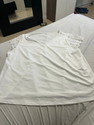 Vit ärmlös topp med spetsdetaljer - Säljer en vit ärmlös topp med v-ringning och fina spetsdetaljer på axlarna. Toppen har en lös passform och är tillverkad i ett mjukt material som känns skönt mot huden. Perfekt för en fräsch och enkel look.