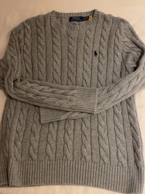 Grå kabelstickad tröja Polo Ralph Lauren - Snygg grå kabelstickad tröja från Polo Ralph Lauren med rund halsringning och diskret logga broderad på bröstet. Tröjan har långa ärmar och ribbade muddar vid ärmslut och nederkant. Perfekt för lager på lager och klassisk stil.
