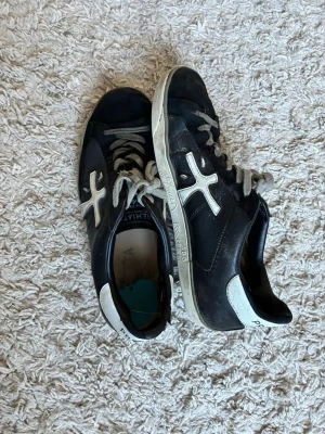 Premiata svarta sneakers med vit detalj - Snygga svarta sneakers från Premiata med vita detaljer och logga på sidan. Skorna har vita snören och en vit sula med text. Baksidan pryds av 'Premiata' och 'Print Edition'. Klassisk låg modell som passar till det mesta.