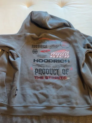 Grå hoodie från Hoodrich med tryck - Säljer en grå hoodie från Hoodrich med stor huva och dragkedja. Framsidan har Hoodrich-logga broderad i rött och svart, medan baksidan är täckt av flera coola tryck och texter i svart, rött och vitt. Hoodien har fickor framtill och är gjord i mjukt sweatshirtmaterial.