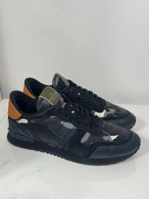 Valentino Rockrunners  - Snygga svarta sneakers från Valentino med kamouflagemönster i grått, vitt och svart. Skorna har en låg profil, svarta skosnören och en kontrasterande brun detalj på hälen. Materialet är en mix av läder och textil, vilket ger en cool och modern look. Hälarna har lagats med lappar