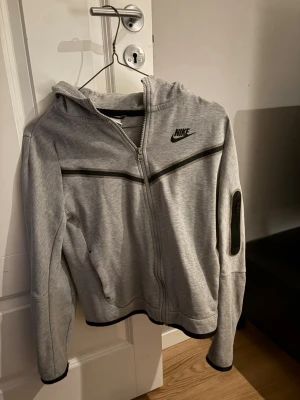 Grå Nike hoodie med dragkedja - Snygg grå hoodie från Nike med svart logga på bröstet och svarta detaljer. Tröjan har hel dragkedja, huva och praktiska fickor samt en extra ficka med dragkedja på ärmen. Perfekt för en sportig och avslappnad stil.