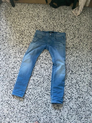 Replay Anbass ljusblå slim jeans - Säljer ett par snygga ljusblå slim fit jeans från Replay, modell Anbass. Jeansen har klassisk femficksdesign, dragkedjegylf och diskreta slitningar för en cool look. Perfekta för dig som gillar en smal passform och mjukt denimtyg.