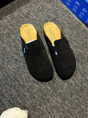Svarta Birkenstock Boston tofflor - Säljer ett par svarta Birkenstock Boston tofflor i mocka med justerbart spänne och klassisk korksula. Insidan är mjuk och bekväm, perfekt för chill dagar hemma eller när du vill ha en clean look. Ikonisk design som alltid är rätt.