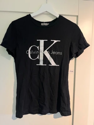 Svart Calvin Klein t-shirt - Svart t-shirt från Calvin Klein Jeans med stort vitt CK-tryck på bröstet. Klassisk rund halsringning och korta ärmar. T-shirten har en slim passform och är tillverkad i mjuk bomull, perfekt för en stilren och enkel look.