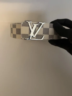 LV - Snyggt bälte från Louis Vuitton med klassiskt rutigt mönster i beige och grått. Bältet har en stor silverfärgad LV-logga som spänne och är tillverkat i skinn. Perfekt accessoar för att lyfta din outfit.