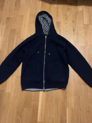Mörkblå som står D I O R hoodie med dragkedja - Mörkblå hoodie med hel dragkedja framtill och justerbar huva. Insidan av huvan har ett mönstrat foder i grått och svart. Klassisk passform och ribbade muddar vid ärmslut och nederkant. Perfekt för en avslappnad streetwear-look.