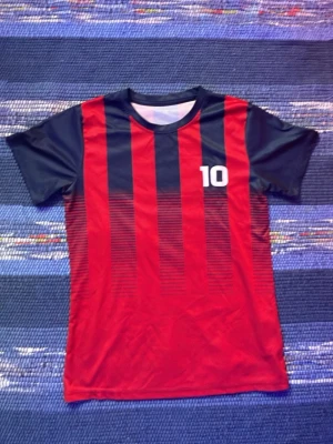 Randig röd och blå träningströja #10 - Sportig t-shirt med vertikala röda och mörkblå ränder samt siffran 10 tryckt på bröstet. Tillverkad i lätt och snabbtorkande polyester, perfekt för fotboll eller träning. Kortärmad modell med klassisk rund hals.