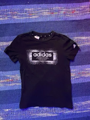 Svart Adidas t-shirt med tryck - Svart t-shirt från Adidas med stort vitt logotryck på bröstet och liten logga på ärmen. Klassisk rund hals och korta ärmar. Perfekt för en sportig och avslappnad stil.