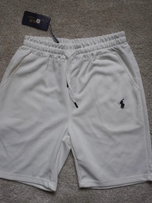 Polo Ralph Lauren Shorts - Snygga vita shorts från Polo Ralph Lauren med elastisk midja och snörning. Klassisk logga broderad i svart på vänster ben. Tillverkade i mjuk bomull för en bekväm känsla. Perfekta för sommardagar och avslappnade outfits. Storleken är XL men känns som L