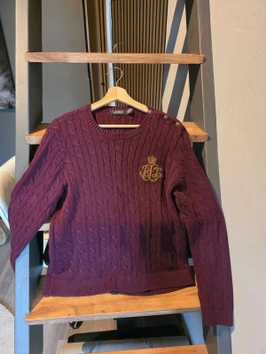 Vinröd kabelstickad tröja Lauren - Vinröd kabelstickad tröja från Lauren Ralph Lauren med rund halsringning, dekorativa guldknappar på axeln och ett broderat emblem på bröstet. Tröjan har långa ärmar och ribbade muddar. Perfekt för dig som gillar klassisk preppy stil. Liten i storlek passade mig när jag hade L i kläder