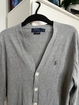 Ralph Lauren Cardigan - Klassisk grå cardigan från Polo Ralph Lauren med v-ringning och vita knappar framtill. Tröjan har lång ärm och diskret broderad logga på bröstet. Tillverkad i mjukt material som känns skönt mot huden. Perfekt att slänga över en t-shirt. Tröjan är i storlek S/M