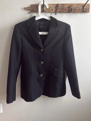 Svart kavaj med guldknappar Ryttarkavaj - Stilren svart kavaj från Equipage med klassisk krage och tre guldknappar framtill. Två fickor med lock och dekorativa knappar bak. Perfekt för dig som gillar en dressad look. Materialet känns tåligt och har en snygg passform.