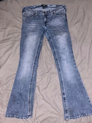Blå bootcut jeans från True Religion - Snygga blå bootcut jeans från True Religion med klassisk femficksdesign och ljusa slitningar på framsidan. Jeansen har broderade bakfickor och en patch med logotyp på bakre linningen. Materialet är denim och passformen är utsvängd nedtill.   Jag haf klippa hå bak för skosnören. Men det synd inte utanpå.    Storlek 29, kolla deras storleksguide