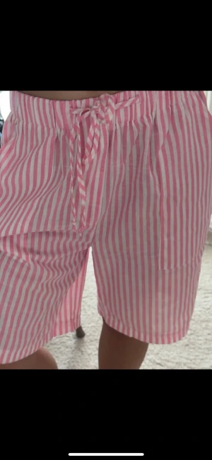 Rosa och vita randiga shorts med snörning - Säljer ett par sköna shorts med rosa och vita ränder och snörning i midjan. De har en avslappnad passform och är perfekta för varma dagar. Shortsen är tillverkade i ett lätt bomullstyg och har en klassisk look.